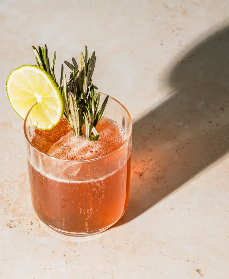 Bellini mocktail served in a glass with a slice of lime & sprigs of rosemary garnish on a light marble surface