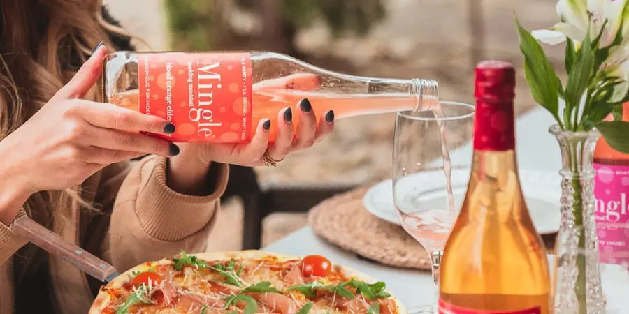 Best alcohol-free cocktail being poured into a wine glass from a Mingle bottle during an elegant outdoor dining experience with pizza.