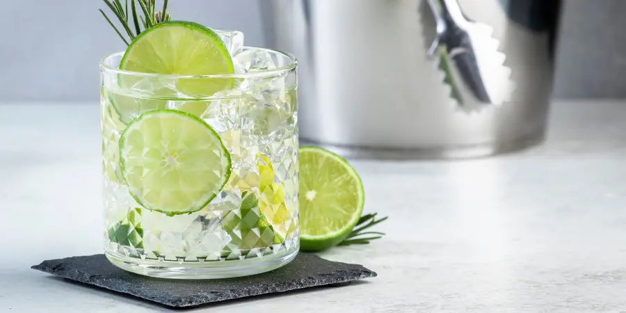 Non-alcoholic gin and tonic in a textured glass with lime slices, rosemary sprigs, and an ice bucket in the background