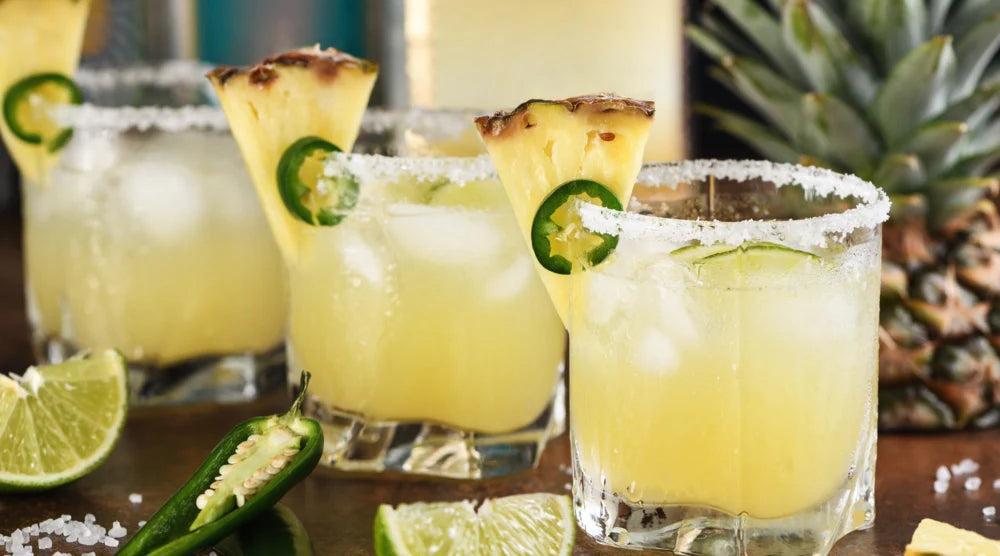 Margarita mocktail served in salt-rimmed glasses, garnished with pineapple slices, jalapeño, & lime wedges, surrounded by fresh ingredients