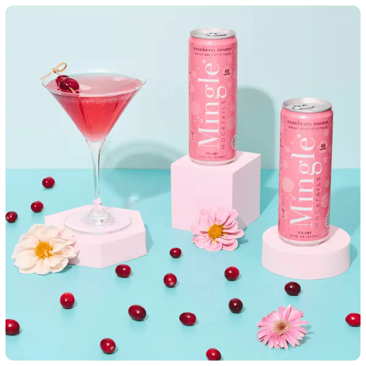 non alcoholic cranberry mocktail being poured into a martini glass, surrounded by cans, cranberries, and flowers on a blue surface