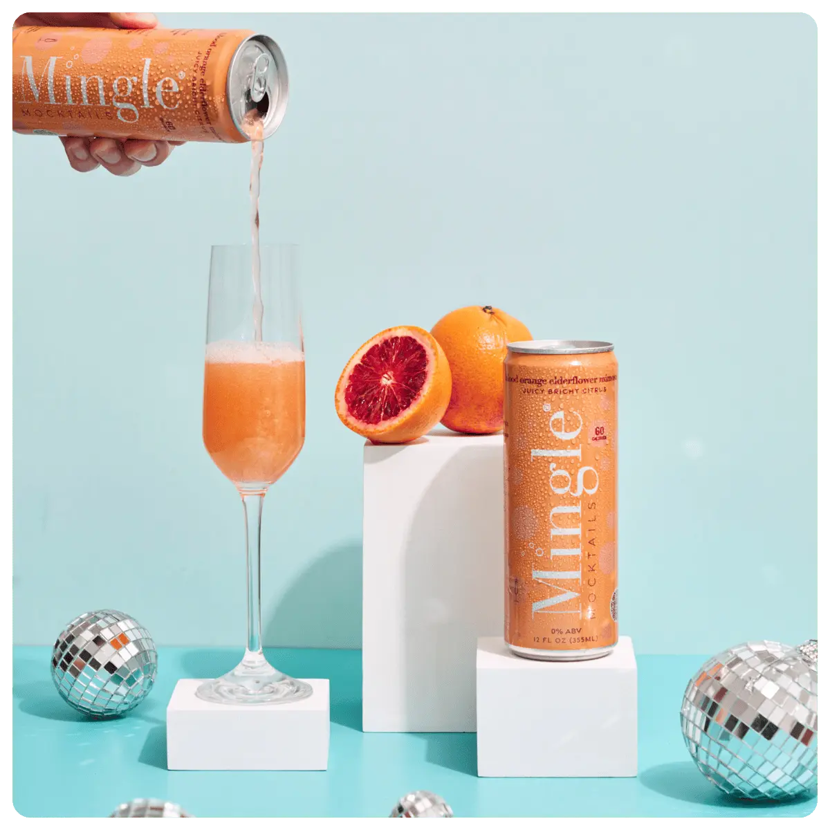 Canned blood orange elderflower mimosa mocktail being poured into a champagne flute beside fresh oranges and mini disco balls.