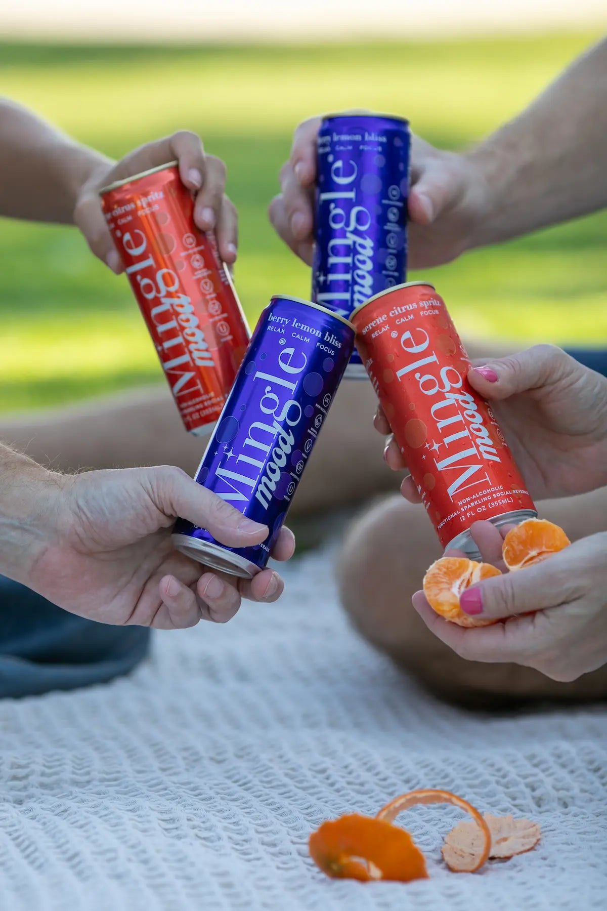 Mocktails with adaptogens in vibrant Mingle Mood cans being shared by friends on a picnic blanket with peeled mandarin oranges.