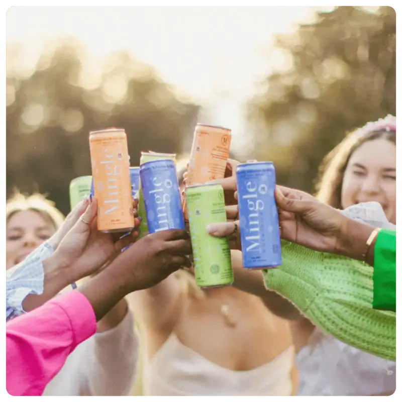 Group of friends raising and clinking cans of Mingle Mocktails in celebration outdoors with smiling faces in the background