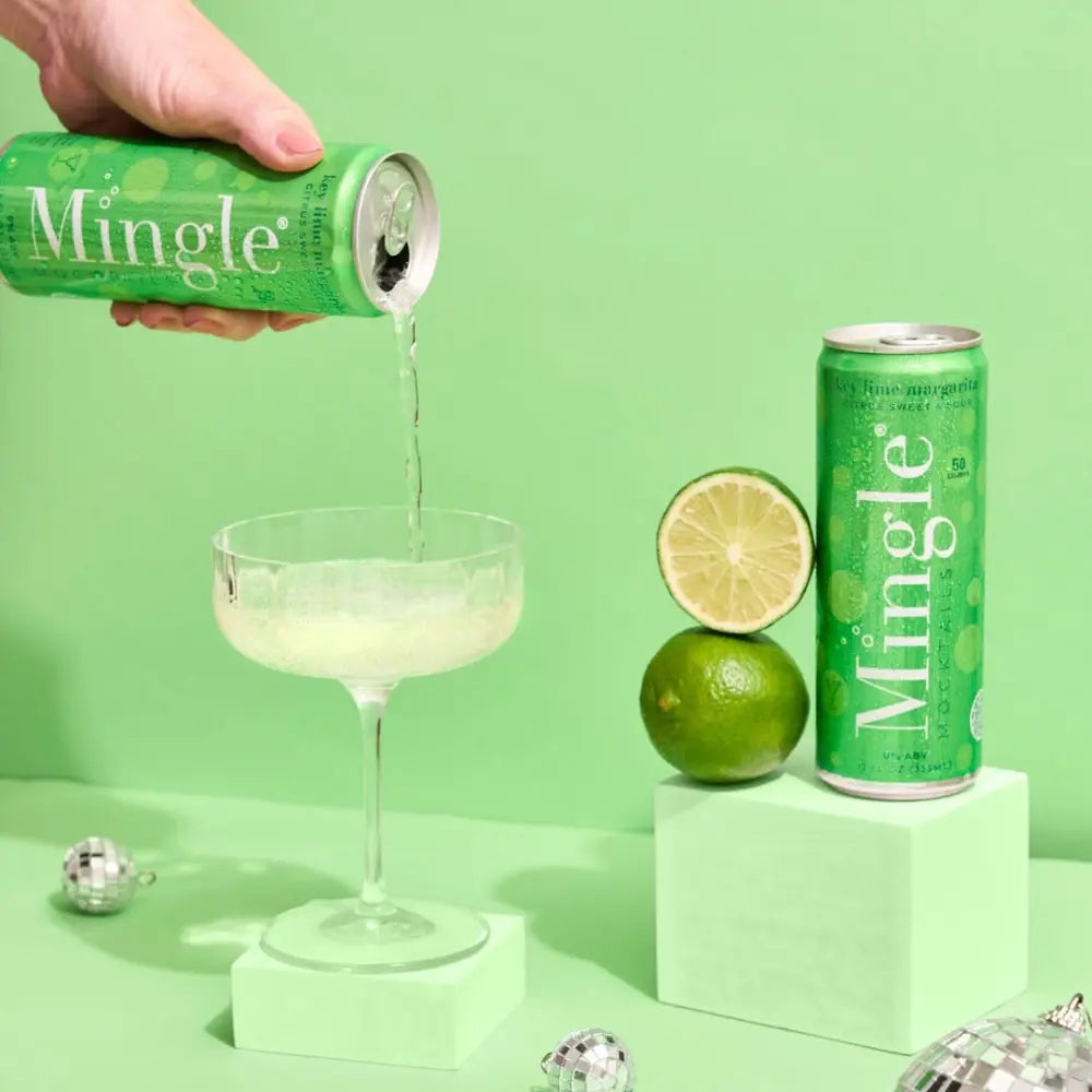 Canned key lime margarita mocktail being poured into a cocktail glass beside fresh limes and a green Mingle can.