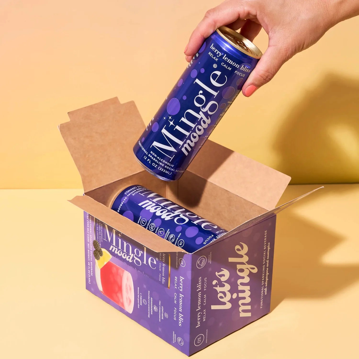 Hand pulling a Mingle Mood Berry Lemon Bliss can from a branded navy blue shipping box on a warm yellow background.