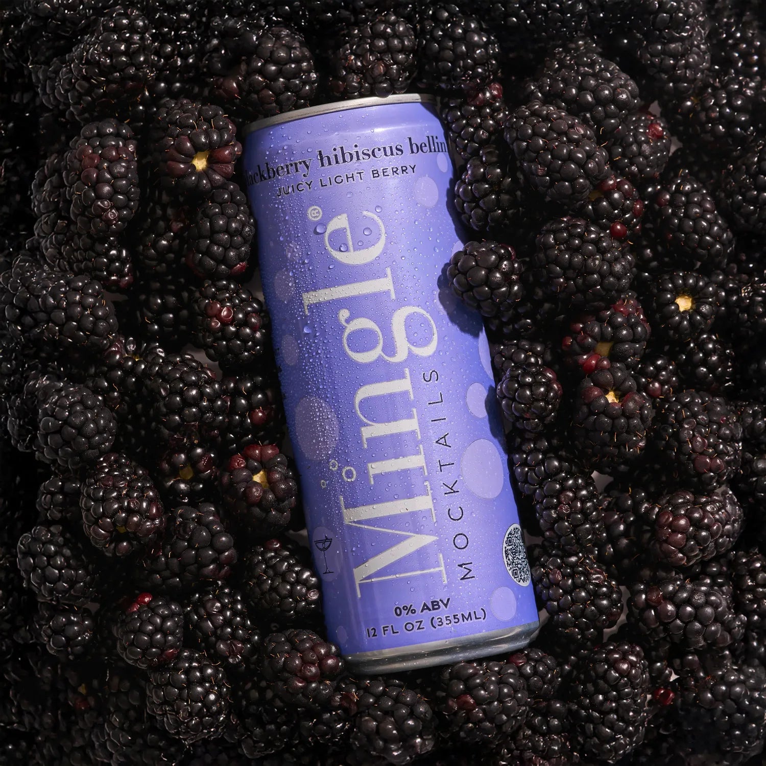 Mingle Mocktails Blackberry Hibiscus Bellini lavender can with condensation resting on a bed of fresh blackberries.