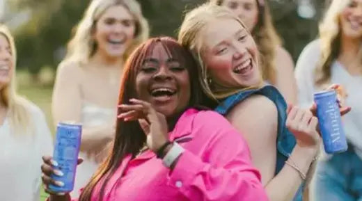Friends laughing & enjoying blackberry hibiscus bellini mocktails outdoors, holding vibrant cans of the drink