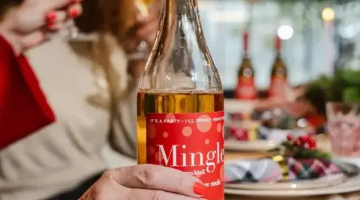 Mocktail bottle labeled "Mingle" held during a festive gathering with decorated table settings in the background