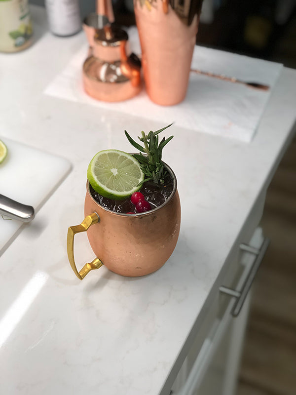 Jingle Berry Mojito mocktail served in a copper mug, garnished with lime, rosemary, and cranberries, on a white countertop with bar tools in the background