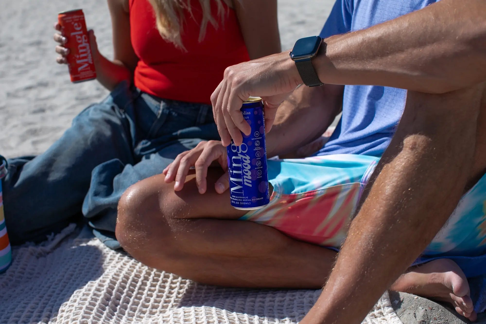 Two people sitting on a blanket on the beach drinking mingle mood berry lemon bliss and mingle mood serene citrus spritz
