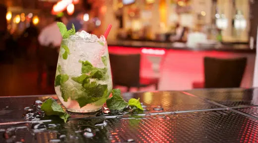 Mojito mocktail served in a glass filled with crushed ice, fresh mint leaves, & a straw, on a glossy bar surface with a vibrant background