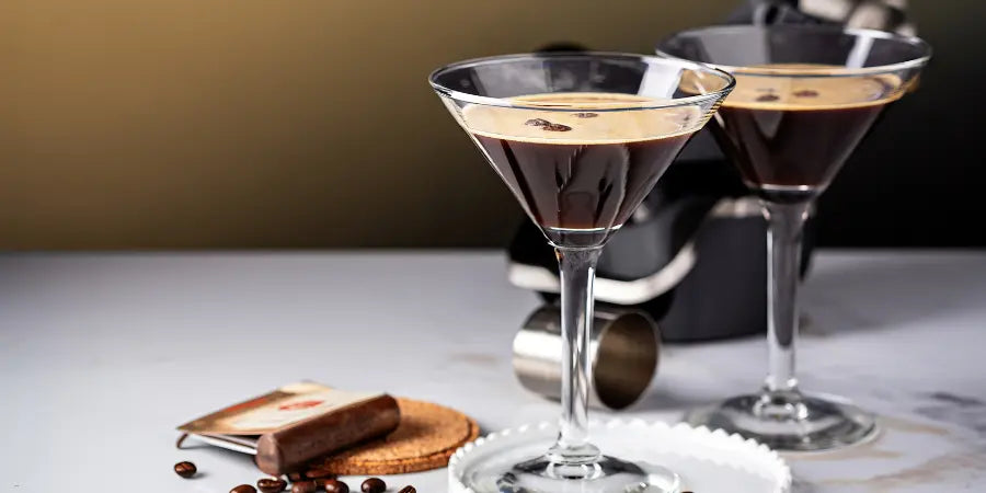 Non-alcoholic espresso martini mocktail in two martini glasses with coffee beans, a biscotti, and a cocktail shaker on a marble surface.