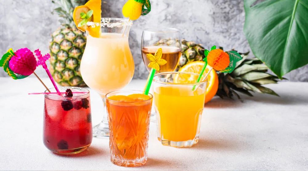 Variety of summer mocktail drinks featuring vibrant non-alcoholic drinks garnished with fresh fruit and colorful straws, set against a tropical backdrop