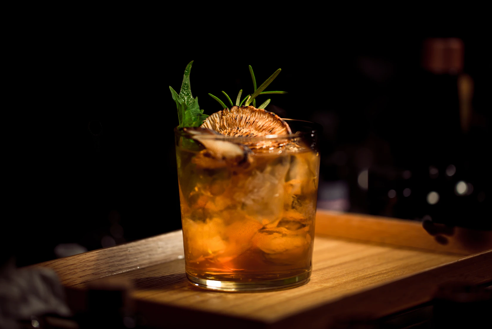 Blood orange elderflower mocktail served in a glass over ice, garnished with a dried orange slice, rosemary, & mint leaves, on a wooden tray