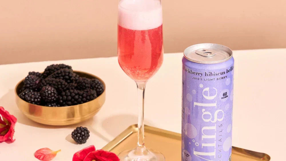 Pink blackberry bellini mocktail in a flute beside a Mingle can, blackberries, and red blooms.