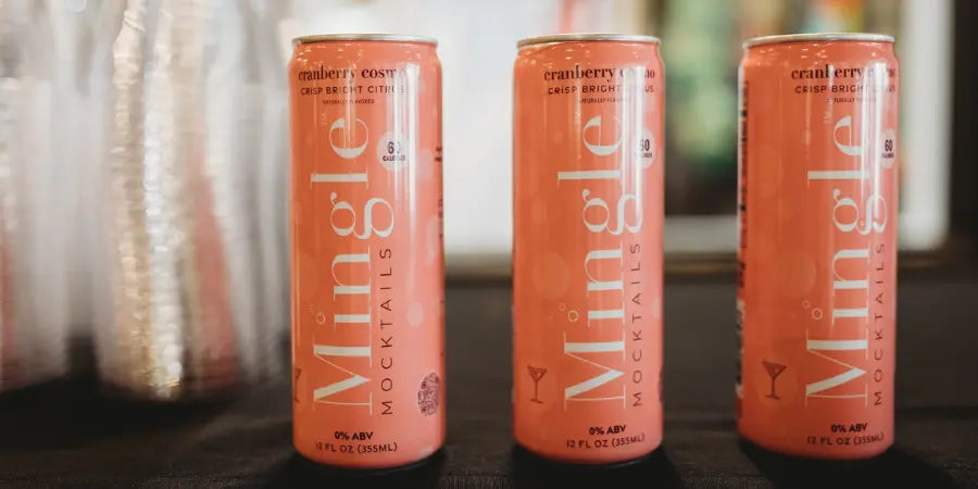 Canned mocktails from Mingle in cranberry cosmo flavor, displayed on a table with clear plastic cups in the background.