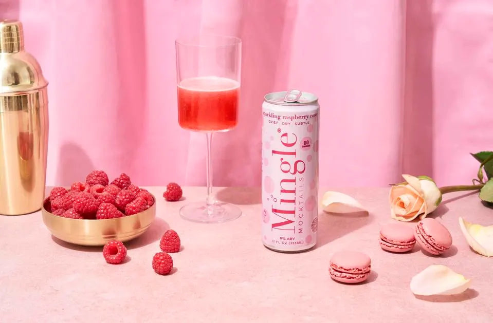 Raspberry rosé mocktail in a glass alongside a can of Mingle, fresh raspberries, & pink macarons