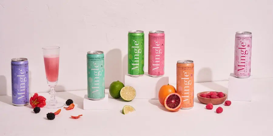 Mingle Mocktails full flavor lineup of colorful cans displayed with fresh fruit including limes, blood orange, and raspberries on a white surface.