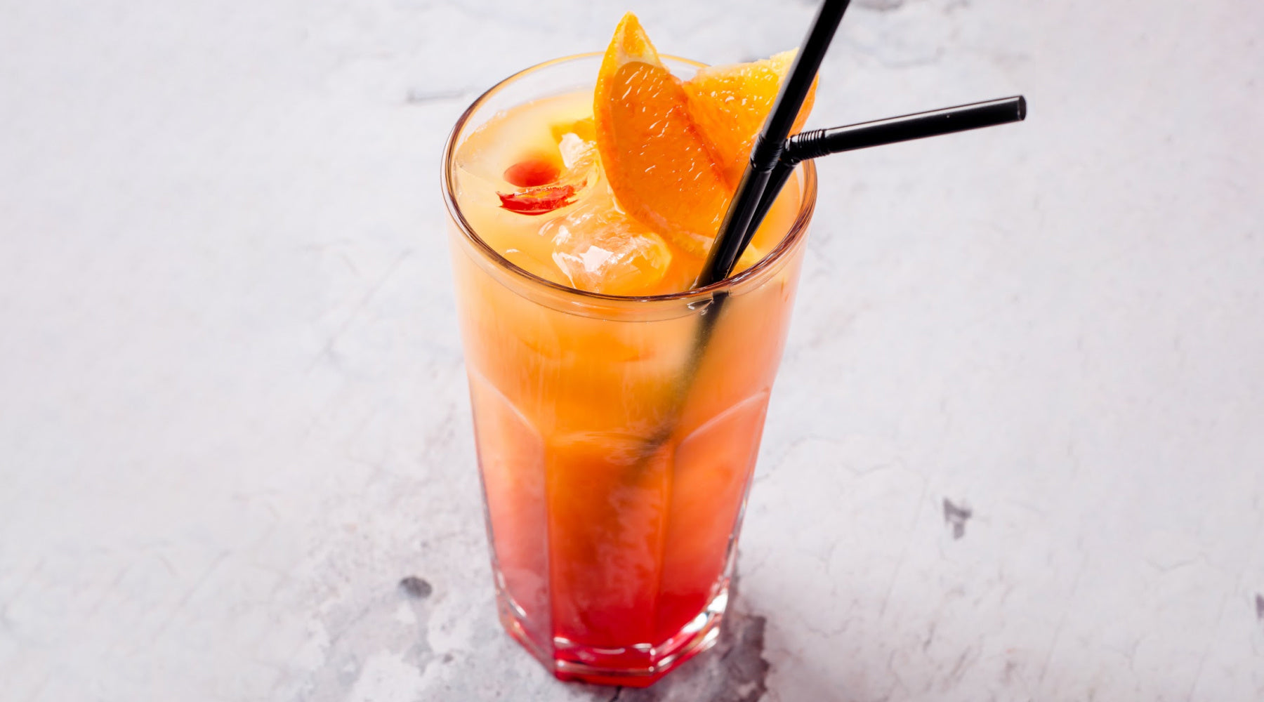 Orange Sunset mocktail served in a tall glass over ice, featuring a gradient of orange & red, garnished with orange slices & a straw