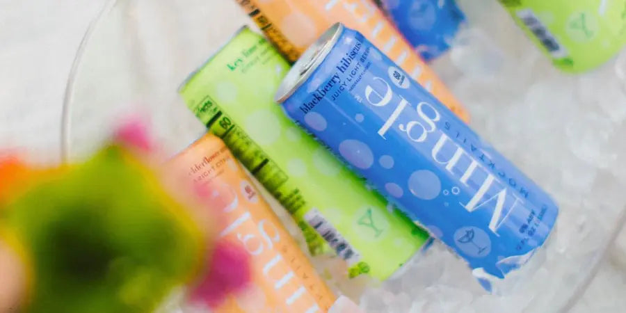 Premade mocktails in colorful Mingle cans chilling on ice, including blackberry hibiscus and key lime flavors, with a blurred cocktail garnish in the foreground