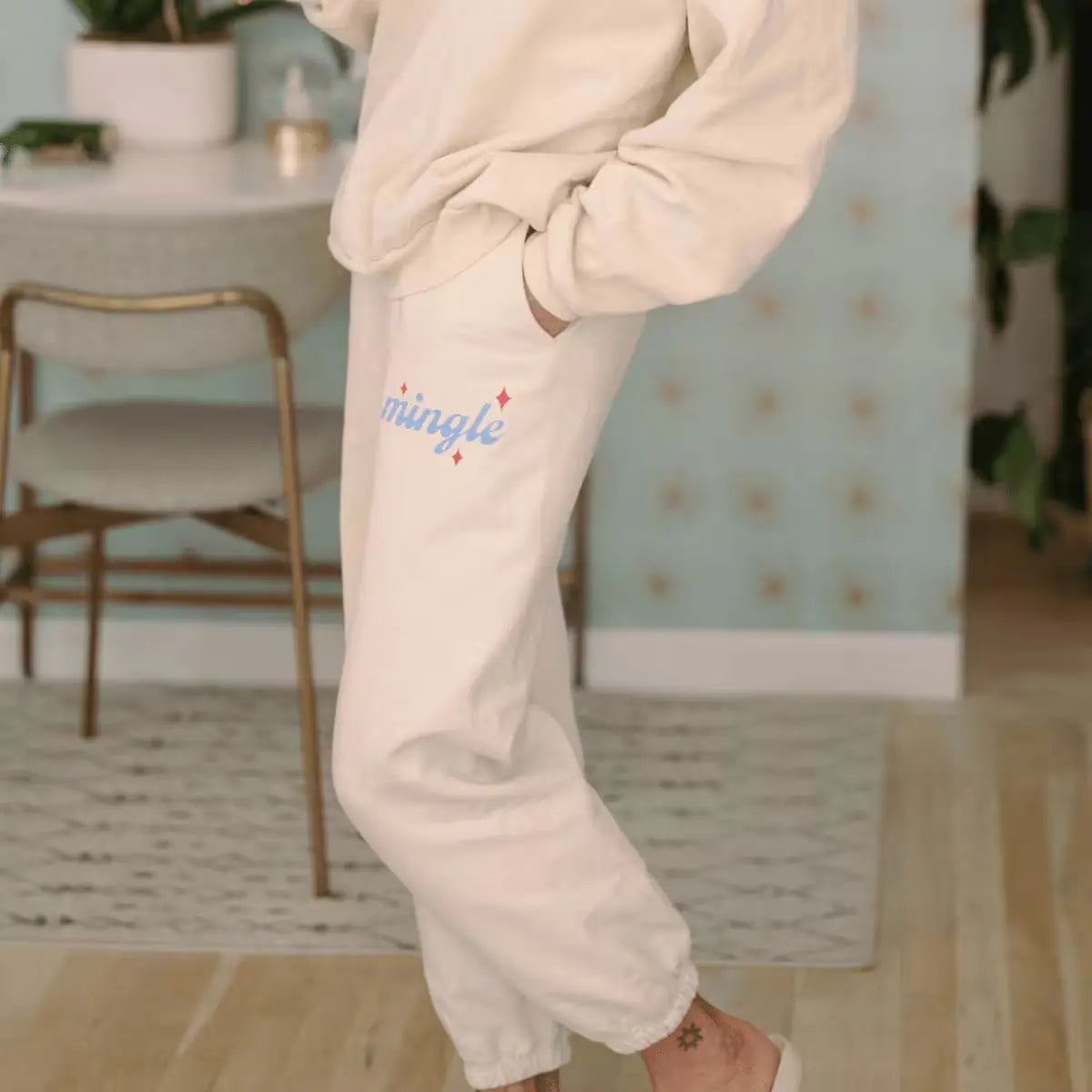 Close-up of a person wearing cream-colored  ‘I Just Wanna Mingle’  sweatpants with "Mingle" branding in blue and red sparkles