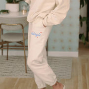 Close-up of a person wearing cream-colored  ‘I Just Wanna Mingle’  sweatpants with "Mingle" branding in blue and red sparkles