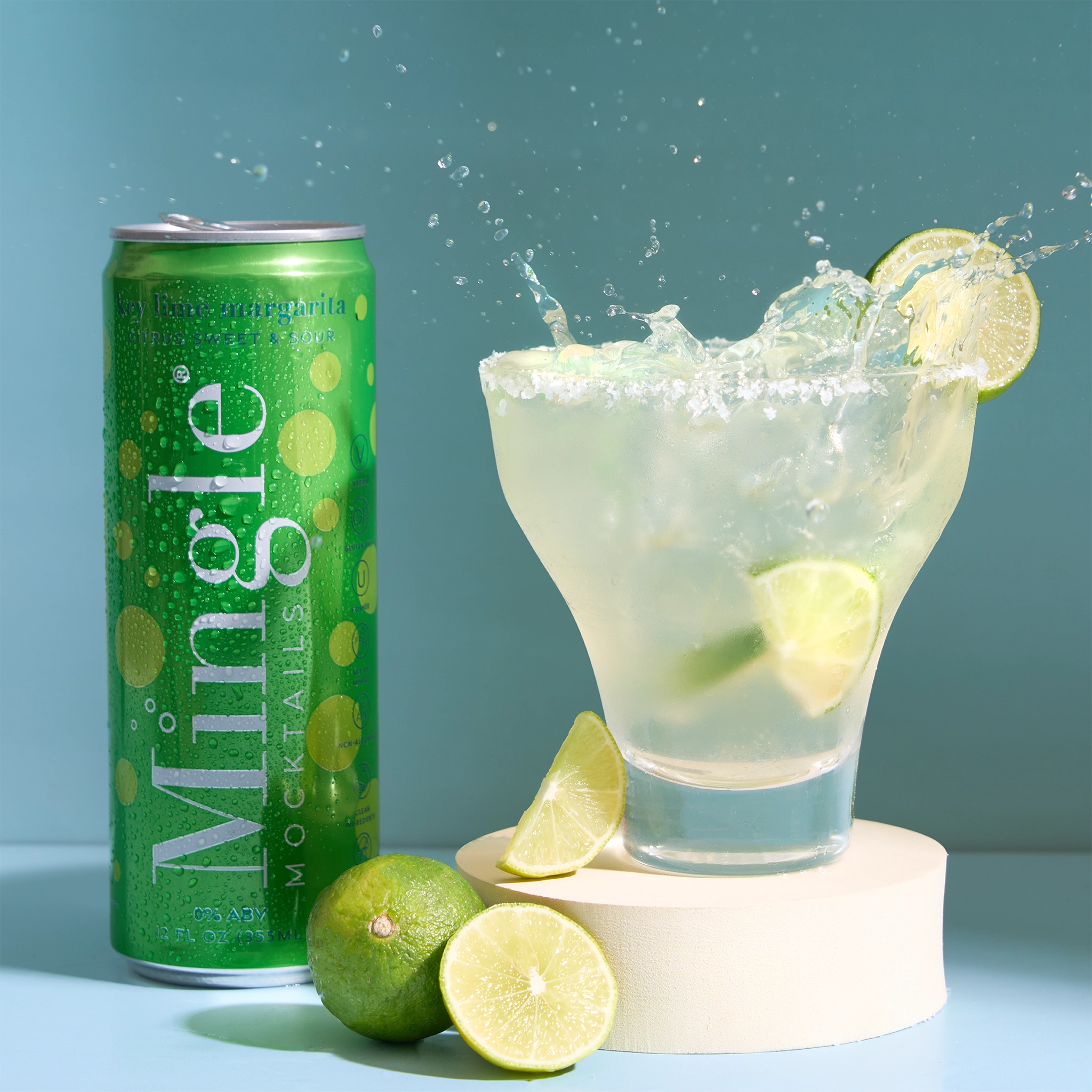 Green Mingle key lime margarita mocktail can beside a splashing margarita glass with lime garnish on a teal background.