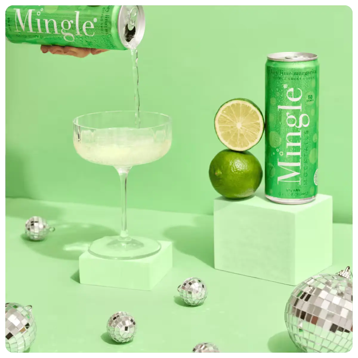 margarita mocktail with lime being poured into a cocktail glass, with a can, lime, and decorative disco balls on a green background