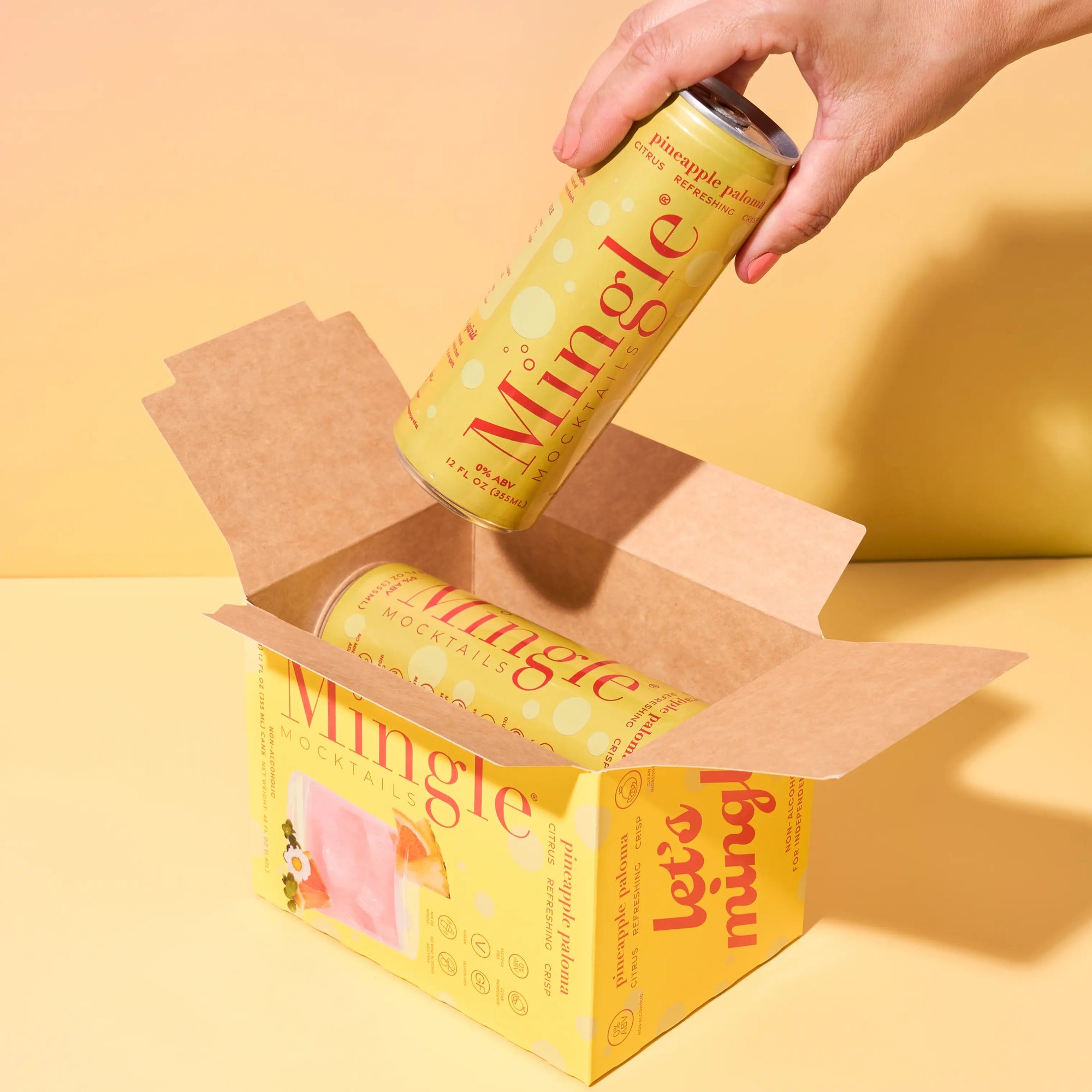Hand pulling a Mingle Mocktails Pineapple Paloma can from a branded yellow shipping box on a warm yellow background.