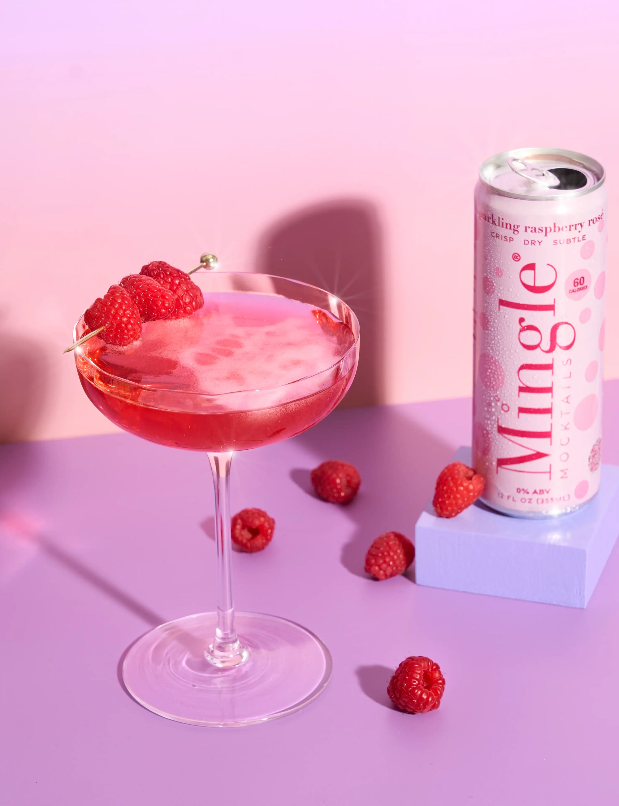 Mingle Mocktails Sparkling Raspberry Rosé served in a coupe glass with raspberry garnish beside the can on a lavender surface.
