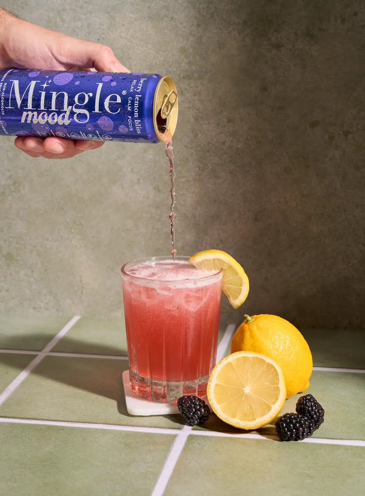 Berry functional mocktail poured from Mingle Mood can into glass with lemon garnish, blackberries, and lemons on green tile