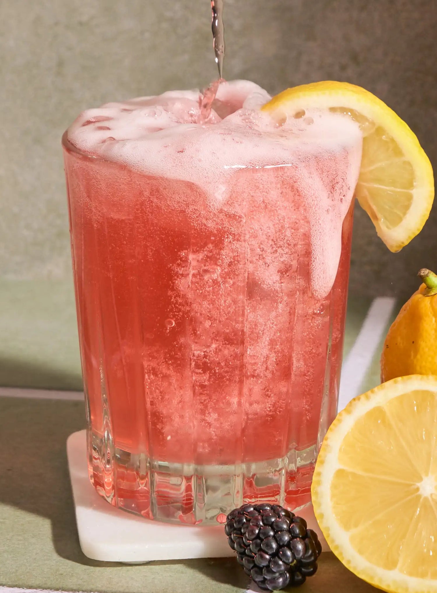 Mingle Mood drink can being poured into a glass with lemons and blackberries on a tiled surface.