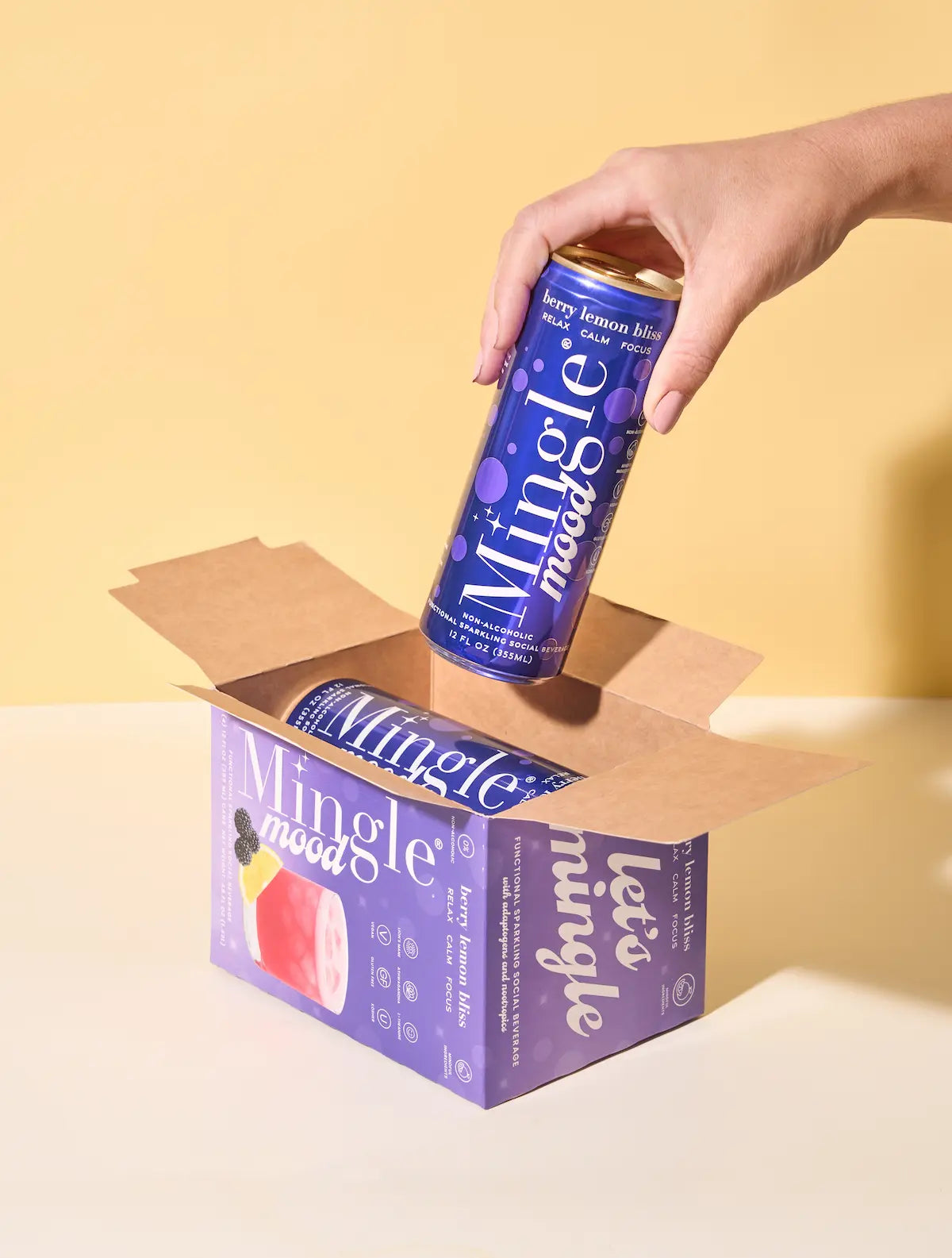 Berry flavored functional mocktail can being lifted from a branded Mingle Mood box against a pastel yellow background.