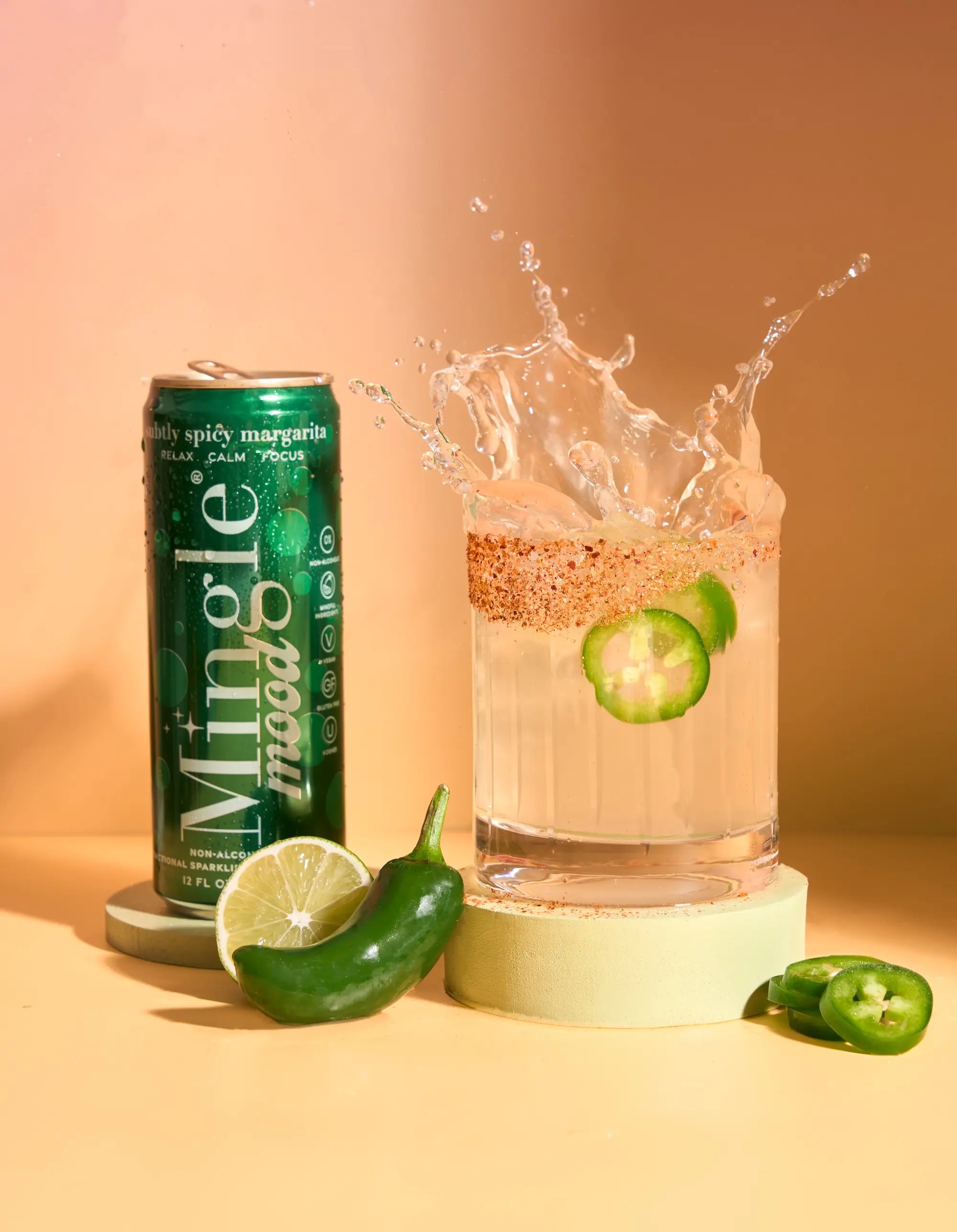 Mingle Mood subtly spicy margarita can beside a jalapeño-rimmed glass mid-splash with lime.