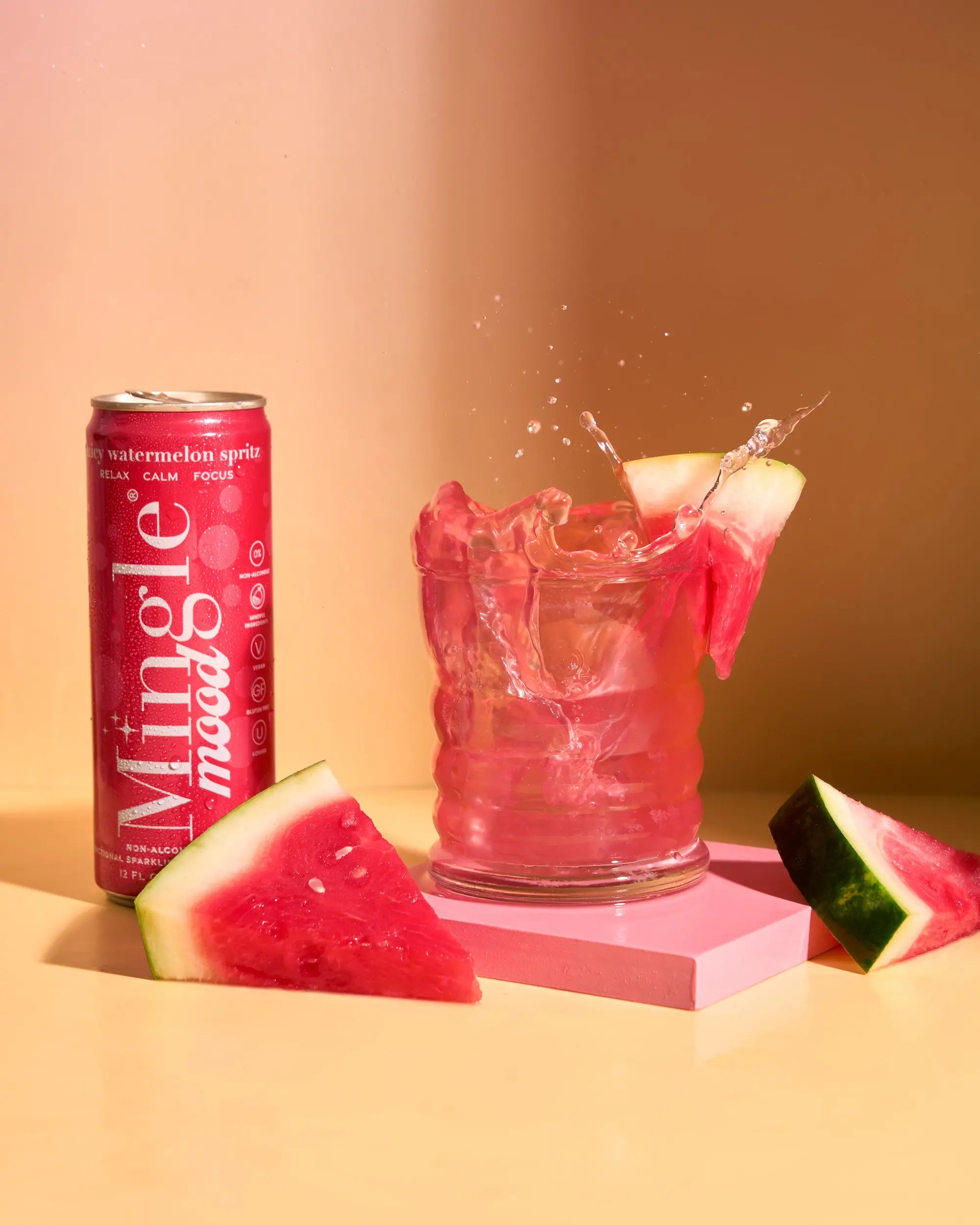 Mingle Mood juicy watermelon spritz can beside a splashing pink mocktail with fresh melon slices.