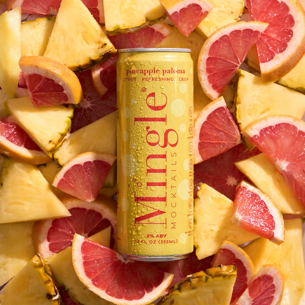 Yellow Mingle Pineapple Paloma non-alcoholic mocktail can nestled among sliced pineapple and pink grapefruit.