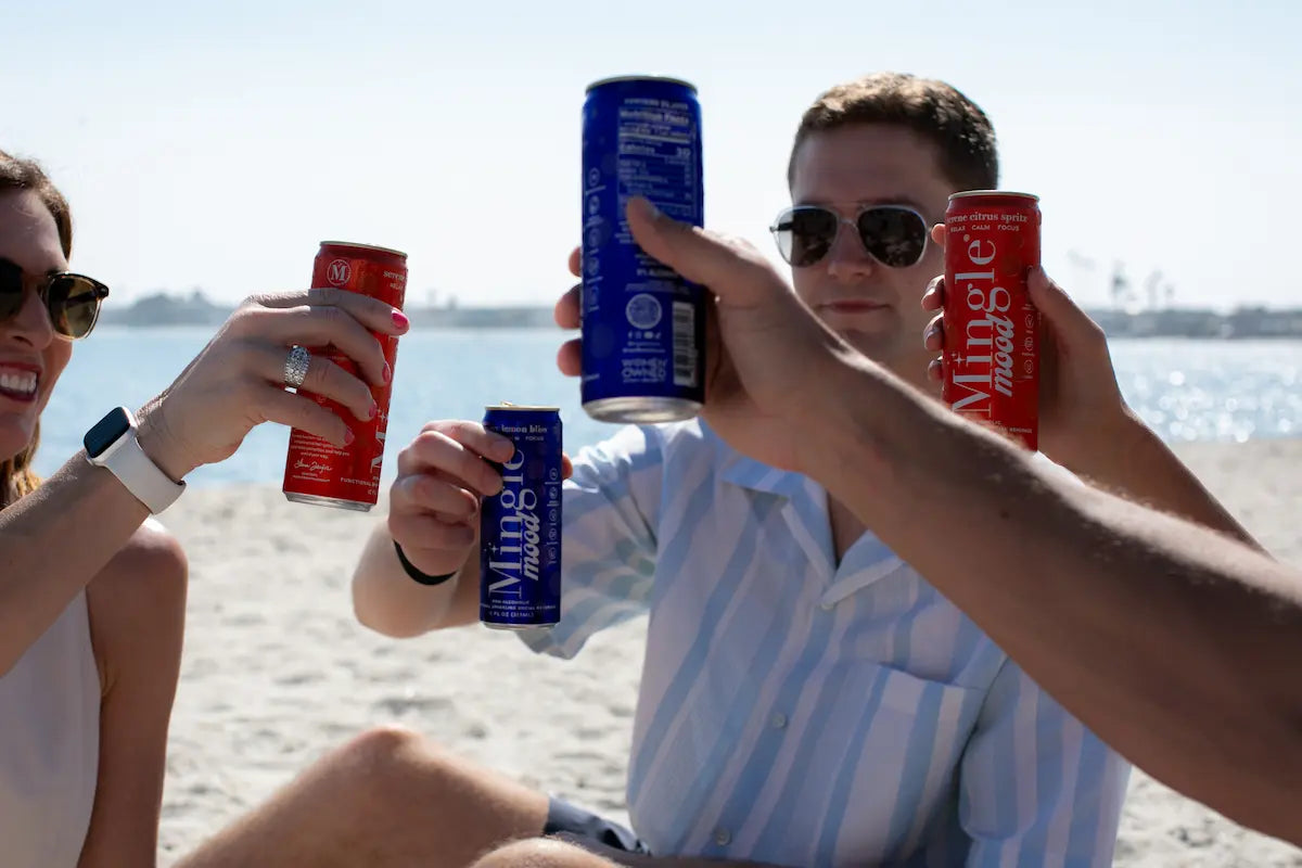 Mocktails with adaptogens in colorful Mingle Mood cans being toasted by friends enjoying a sunny day at the beach.
