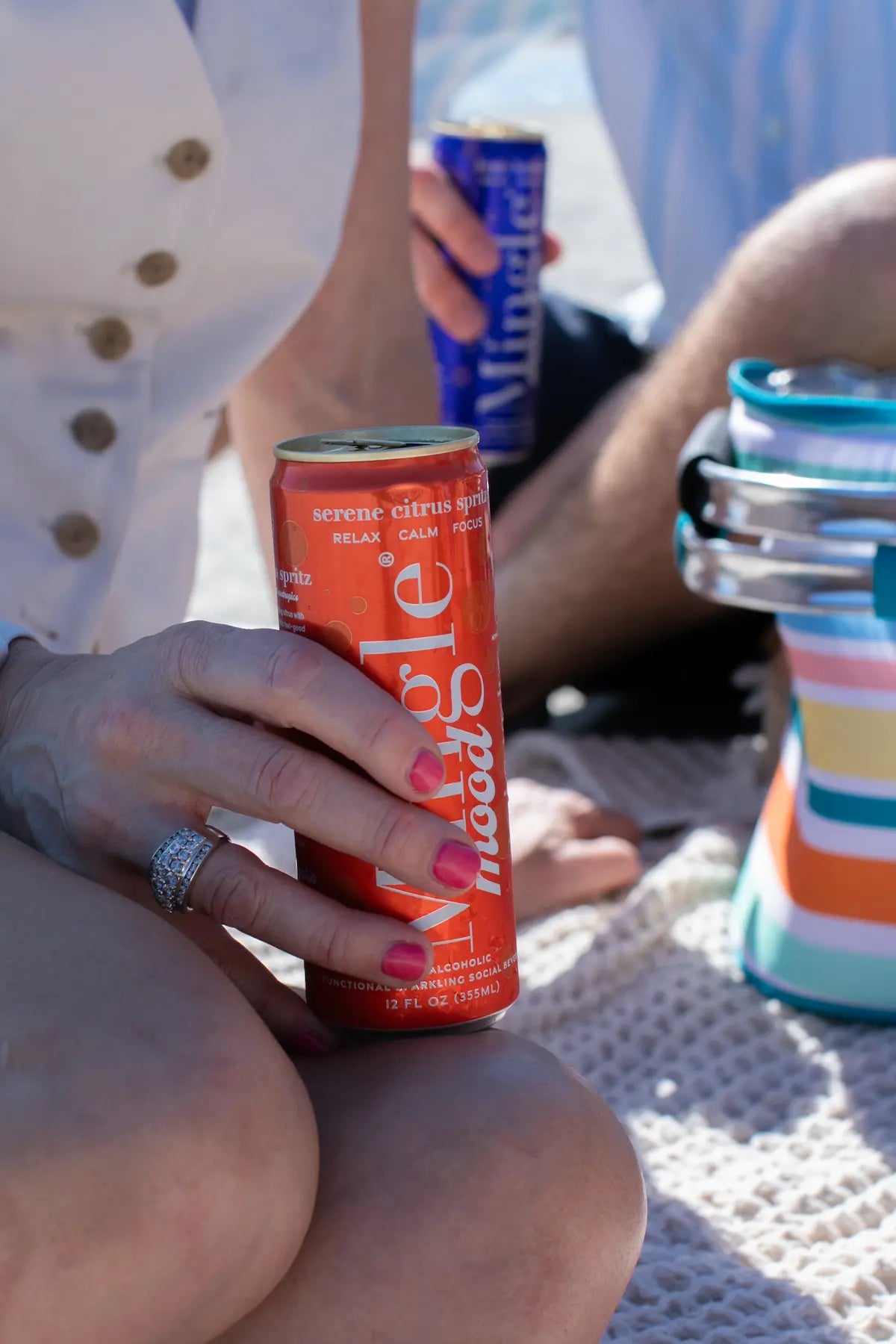 Functional beverage with adaptogens in a citrus-flavored Mingle Mood can held by a woman during a sunny beach picnic