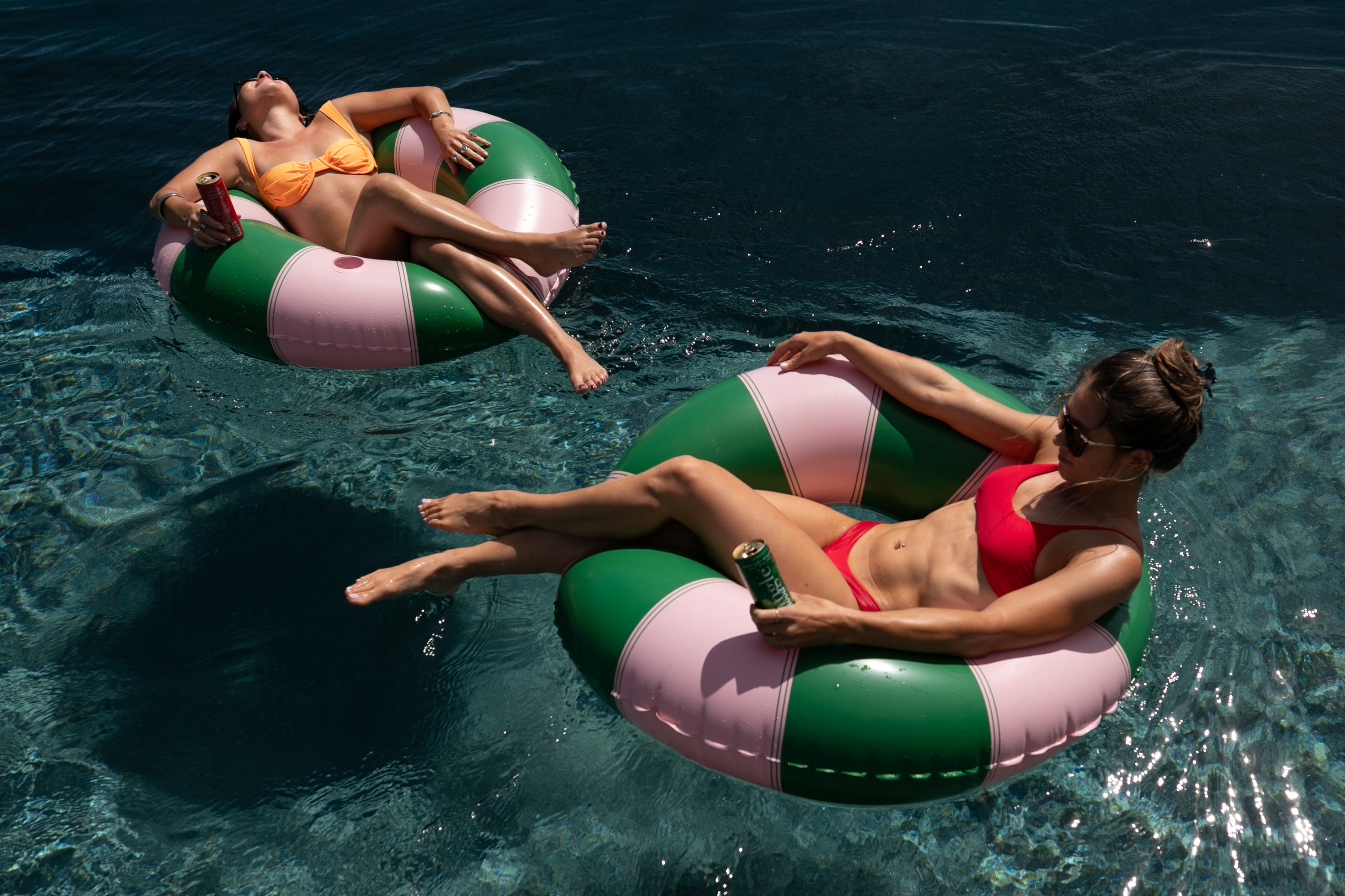 Two friends floating on pink and green pool tubes sipping canned Mingle mocktails in sunny water.