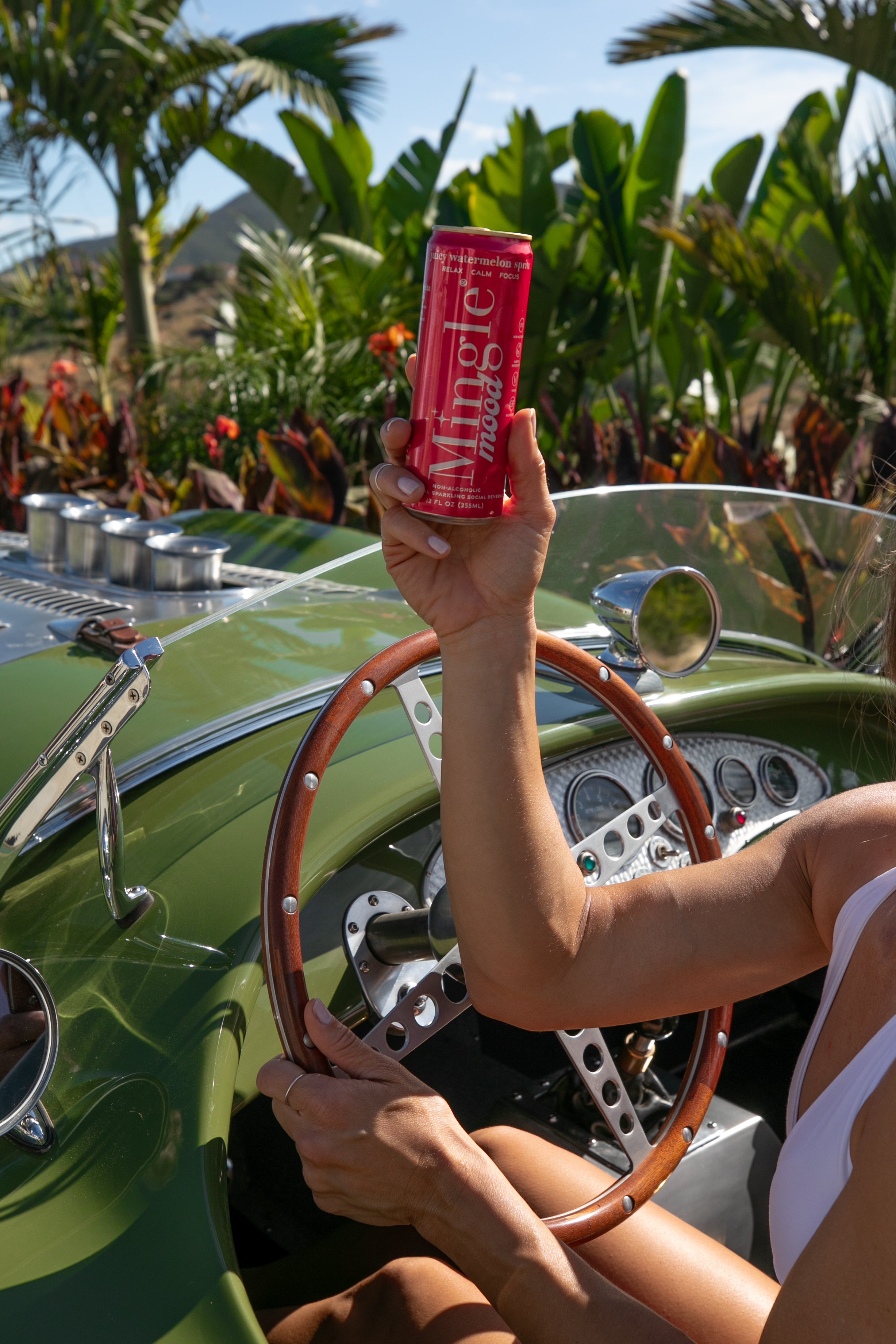 Raised pink Mingle Mood juicy watermelon spritz can held above a vintage green convertible's wooden steering wheel.
