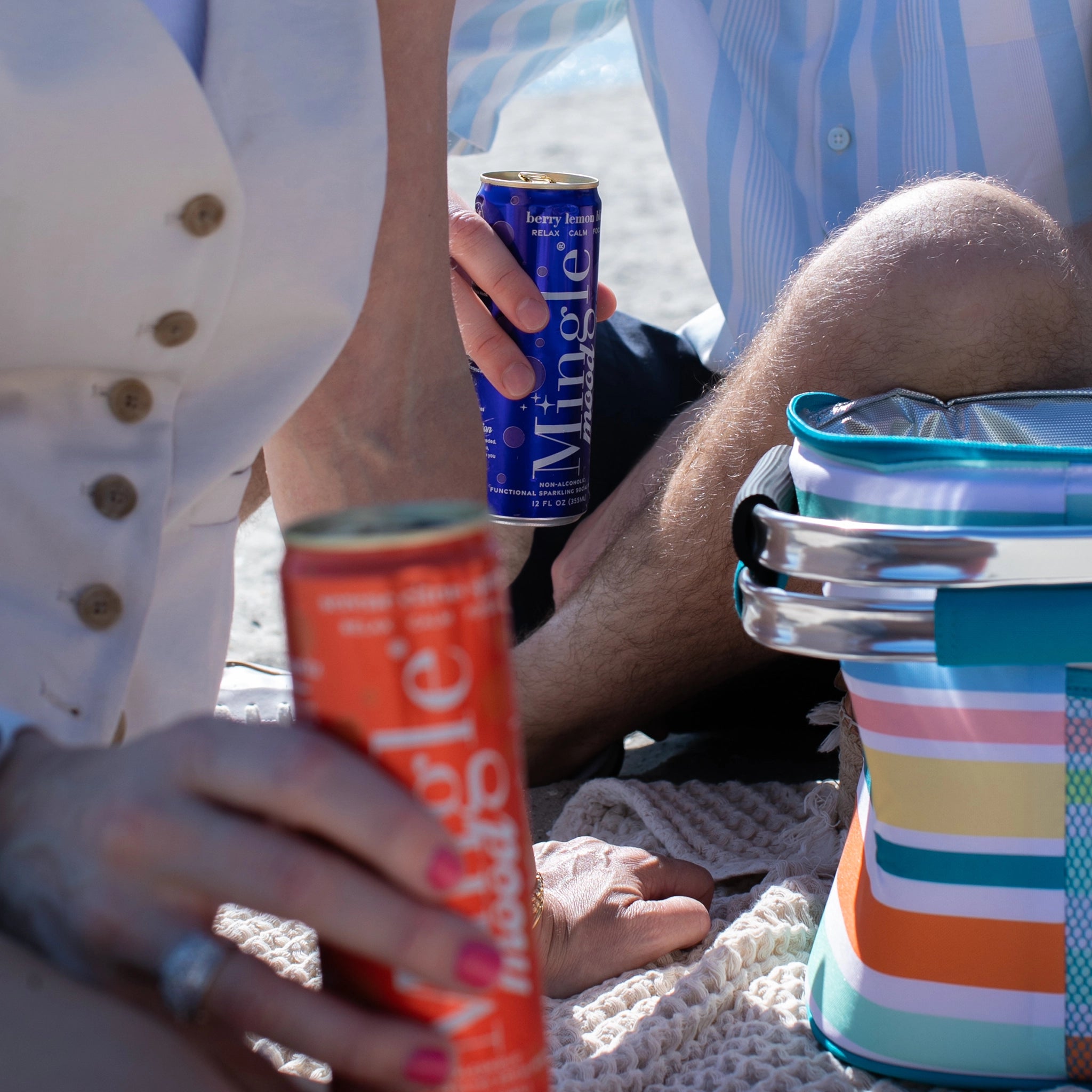 Two people relaxing on a beach blanket holding Mingle Mood Berry Lemon Bliss and Serene Citrus Spritz adaptogen drink cans.