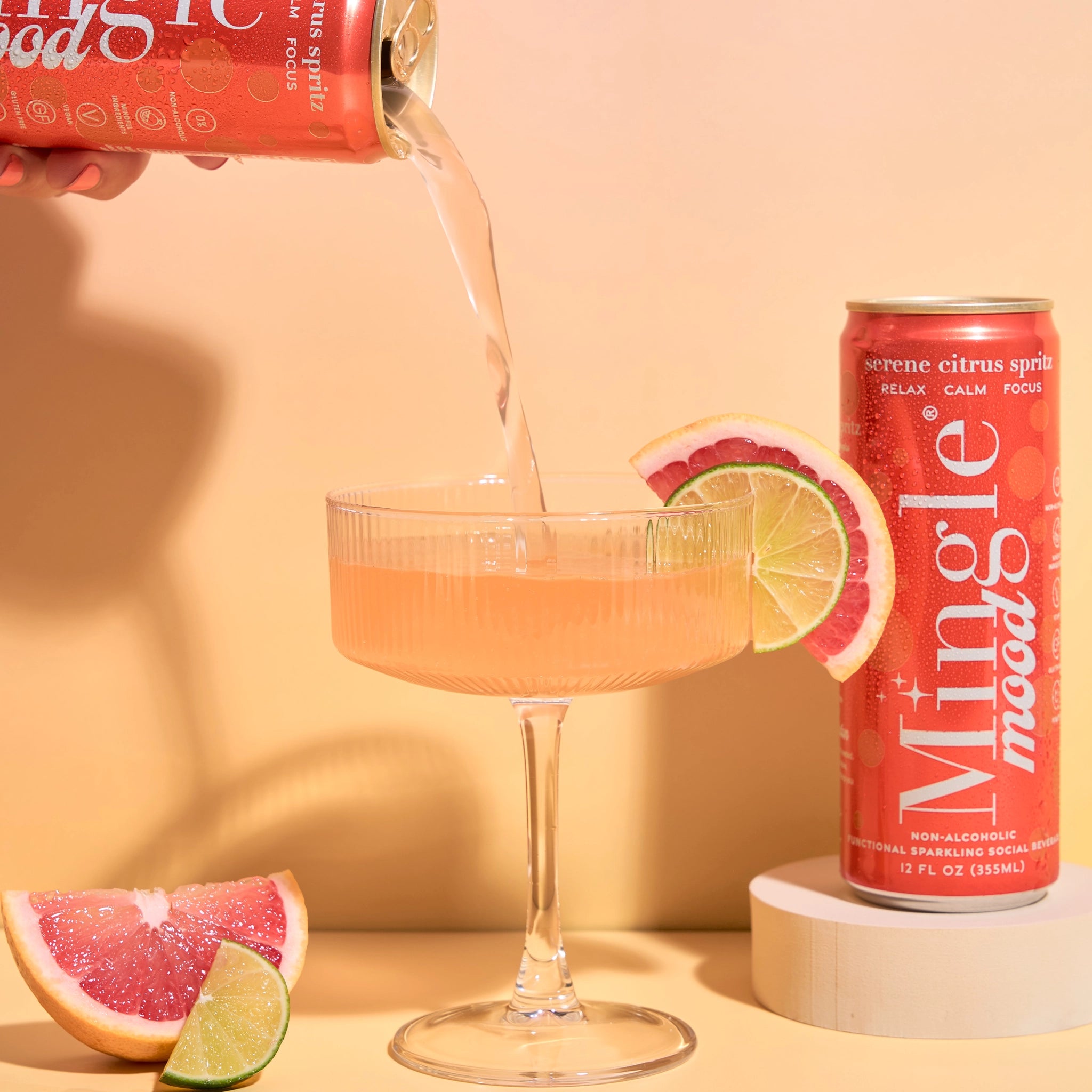 Mingle Mood Serene Citrus Spritz can on a pedestal beside a coupe glass garnished with grapefruit and lime slices.