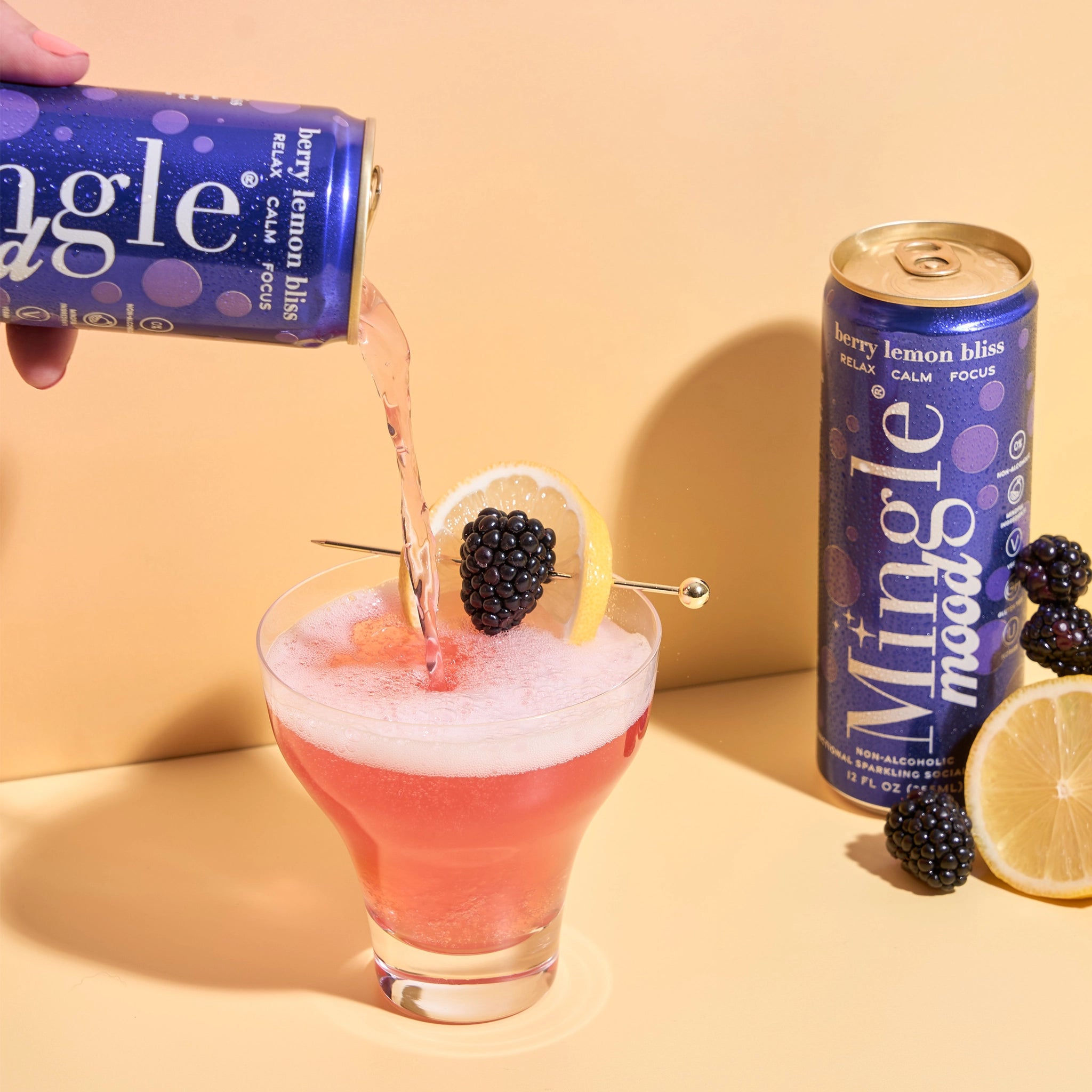 Mingle Mood Berry Lemon Bliss being poured into a garnished glass beside fresh blackberries and lemon halves.