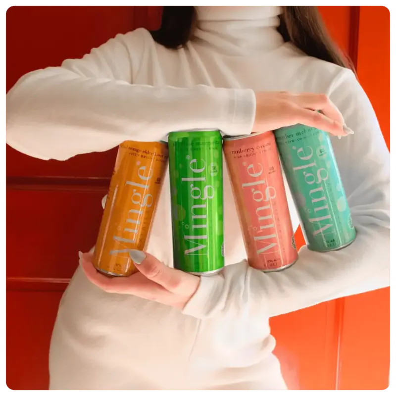 Person in a white turtleneck holding four colorful Mingle Mocktails cans against a vibrant red background