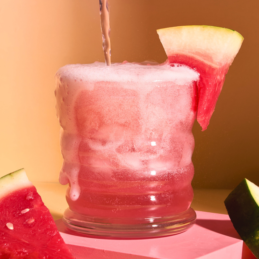 Pink watermelon mocktail bubbling over a wavy glass with fresh watermelon wedges on a peach backdrop.