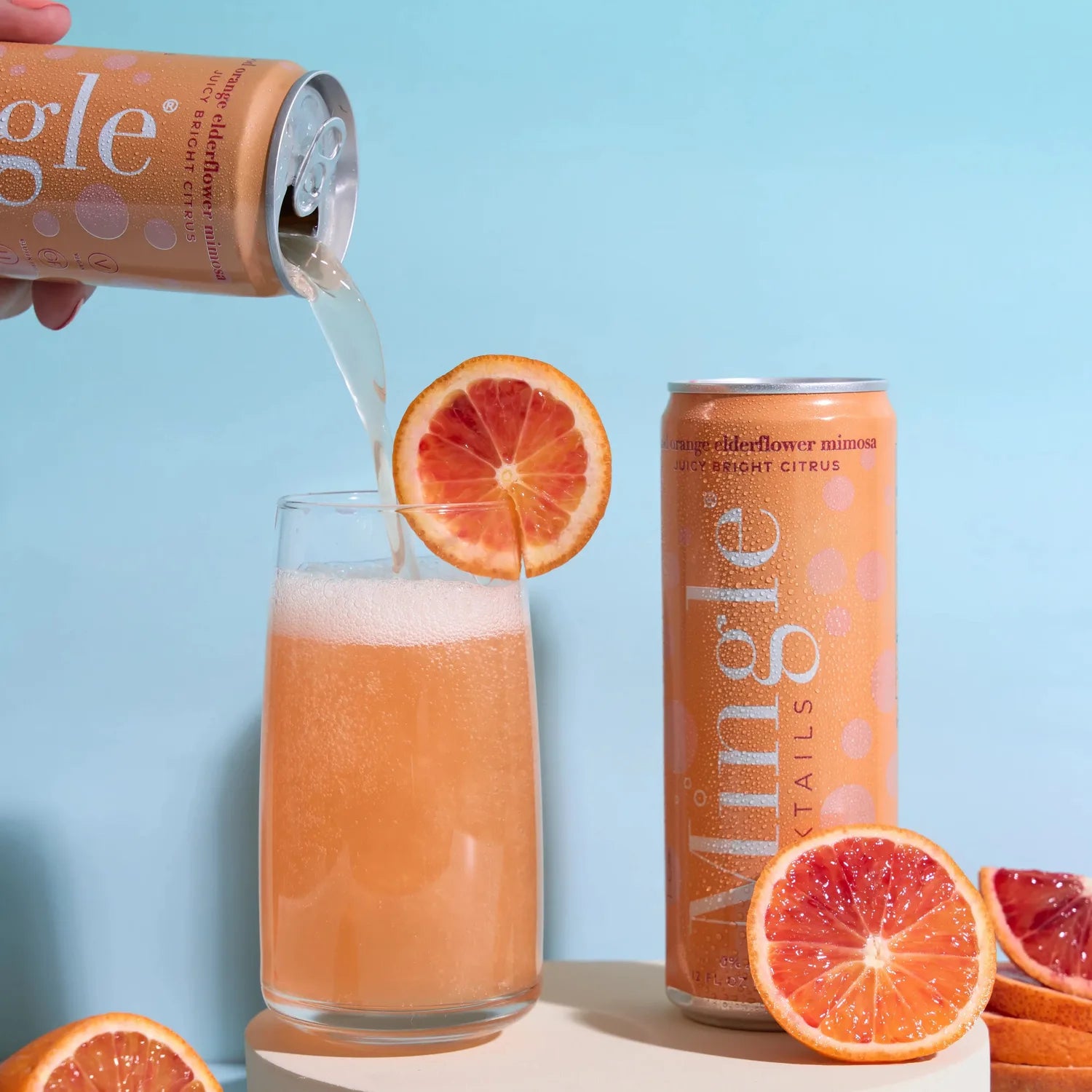 Hand pouring Mingle Mocktails Blood Orange Elderflower Mimosa into a tall glass surrounded by blood orange slices on a blue background.