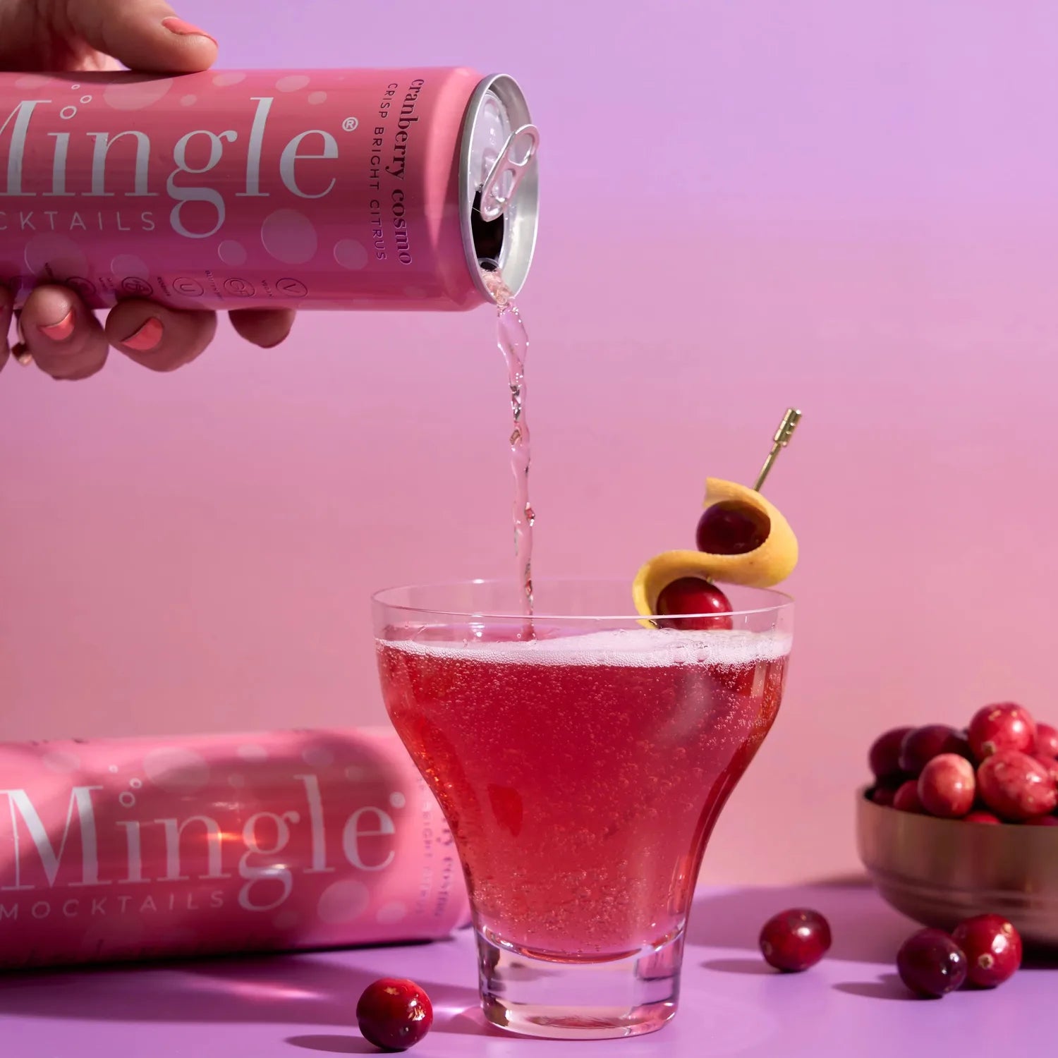 Hand pouring Mingle Mocktails Cranberry Cosmo from a pink can into a cocktail glass garnished with cranberries and lemon twist.
