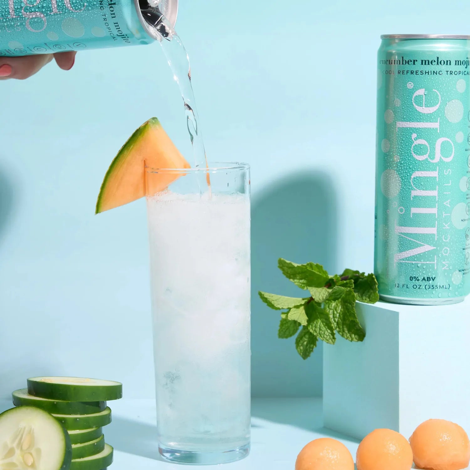 Hand pouring Mingle Mocktails Cucumber Melon Mojito into a tall glass with melon wedge, cucumber slices, and mint on a teal background.