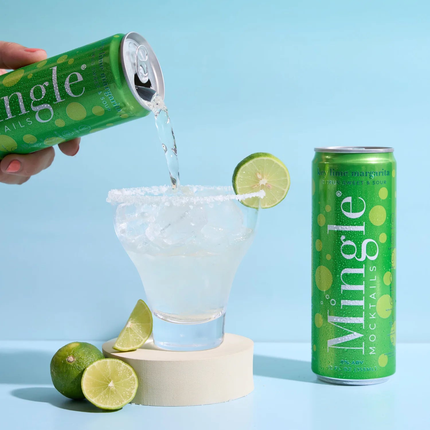 Hand pouring Mingle Mocktails Key Lime Margarita into a salt-rimmed glass with lime wheel beside the green can on a blue background.